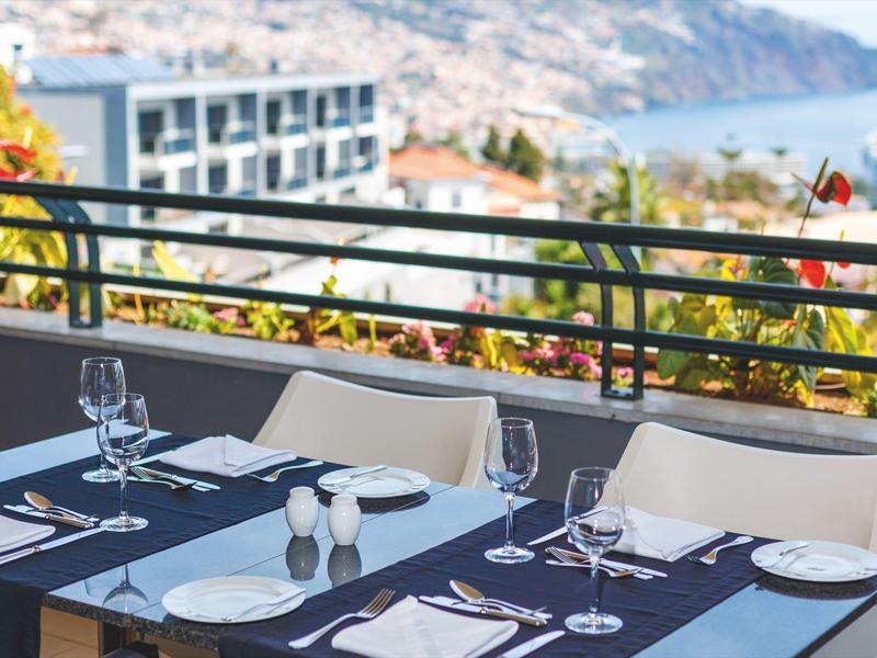 Mesa al aire libre con vajilla y copas, con vista a un edificio y colinas a lo lejos.