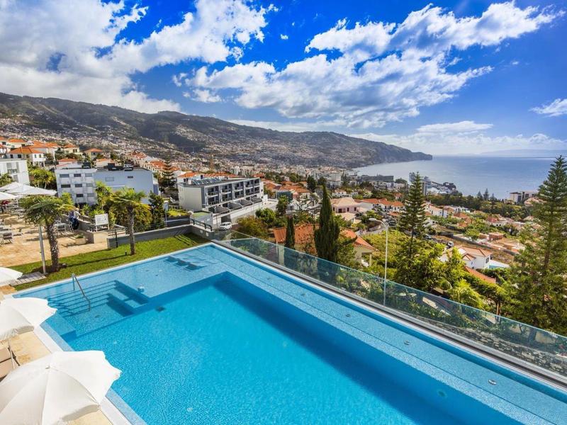 Piscina infinity con vista a la ciudad, la costa y montañas bajo cielo azul con nubes.