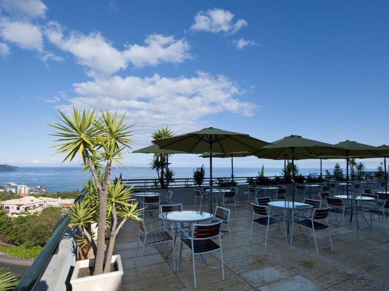 Terraza con mesas y sillas, vista al mar, colinas y ciudad bajo cielo despejado.