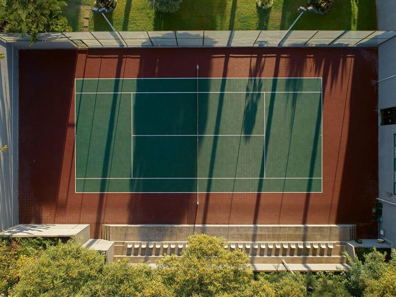 Vista aérea de una cancha de tenis con áreas de juego rojas y verdes rodeada de árboles.