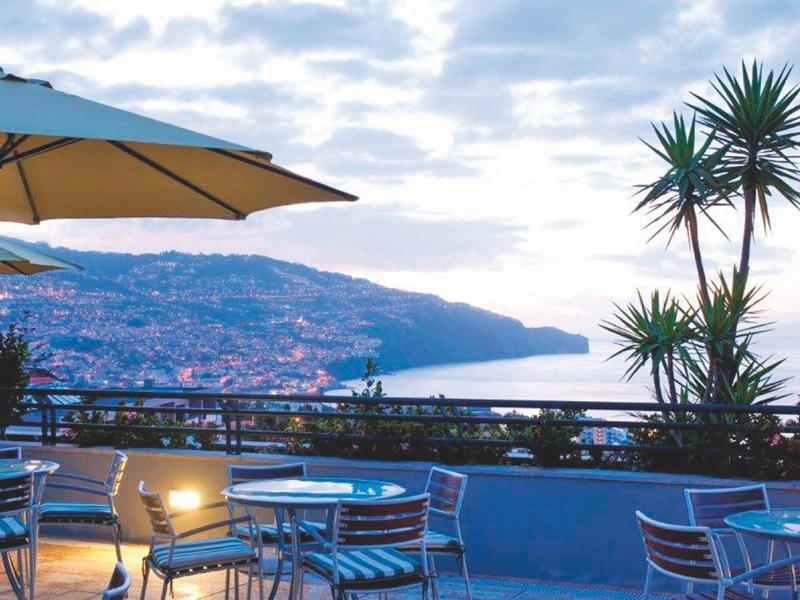 Terraza de un hotel con mesas, sillas y sombrilla con vistas al mar y colinas.