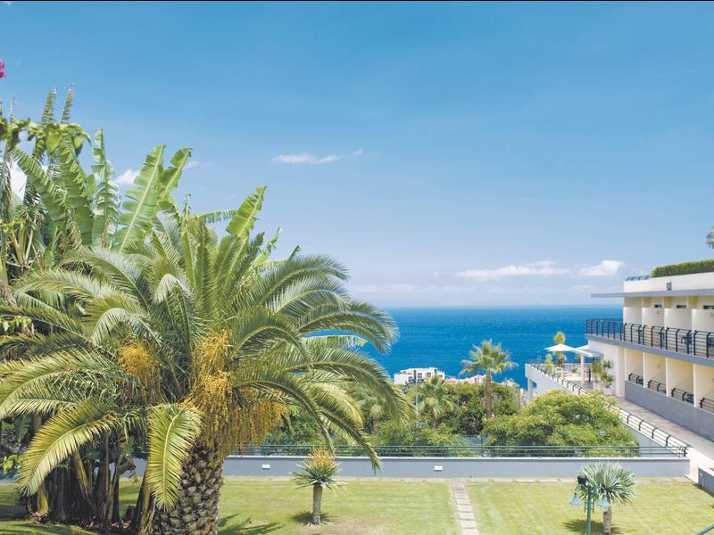 Complejo hotelero con palmeras, flores coloridas y vista al mar azul bajo cielo despejado.