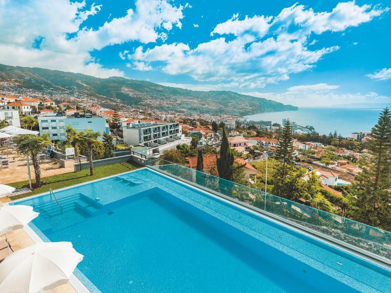 Piscina infinita con vista a la ciudad, montañas y mar en un día soleado.
