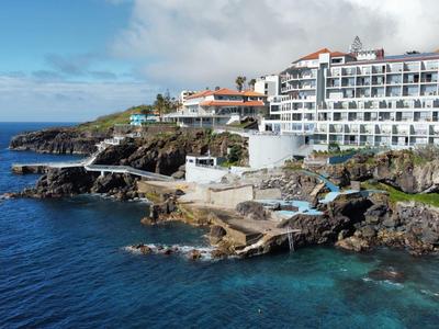 Hotelový komplex na skalnatém pobřeží s bazénem a čistým modrým mořem pod zataženou oblohou.