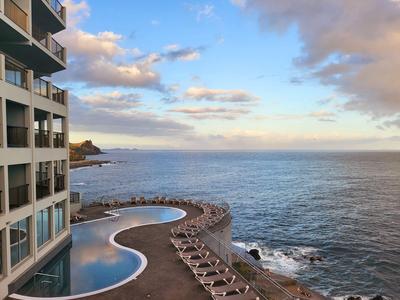 Hotel mit geschwungenem Pool und Liegestühlen am Meer bei Sonnenuntergang.