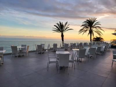 Venkovní restaurace se stoly, židlemi, dvěma palmami a výhledem na moře při západu slunce.