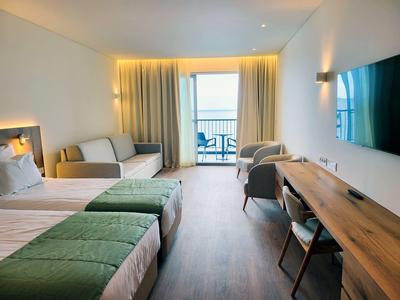 Moderní hotelový pokoj se dvěma jednolůžky, pohovkou, psacím stolem a balkonem s výhledem na moře.