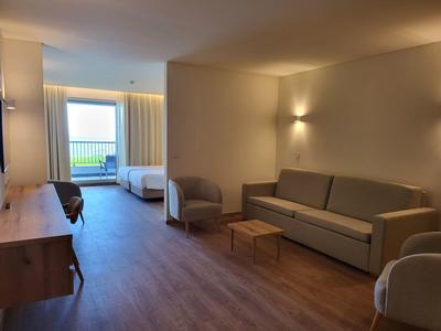 Světle hotelový pokoj se dvěma postelemi, pohovkou, křesly a přístupem na balkon s výhledem ven.