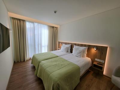 Moderní hotelový pokoj se dvěma jednolůžky, zelenými přikrývkami a velkými okny s závěsy.