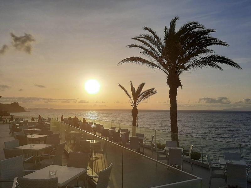 Západ slunce nad mořem s palmami a restaurací na terase u vody.