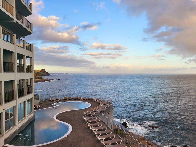 Hotel s bazénovou terasou u moře a lehátky na skalnatém pobřeží.