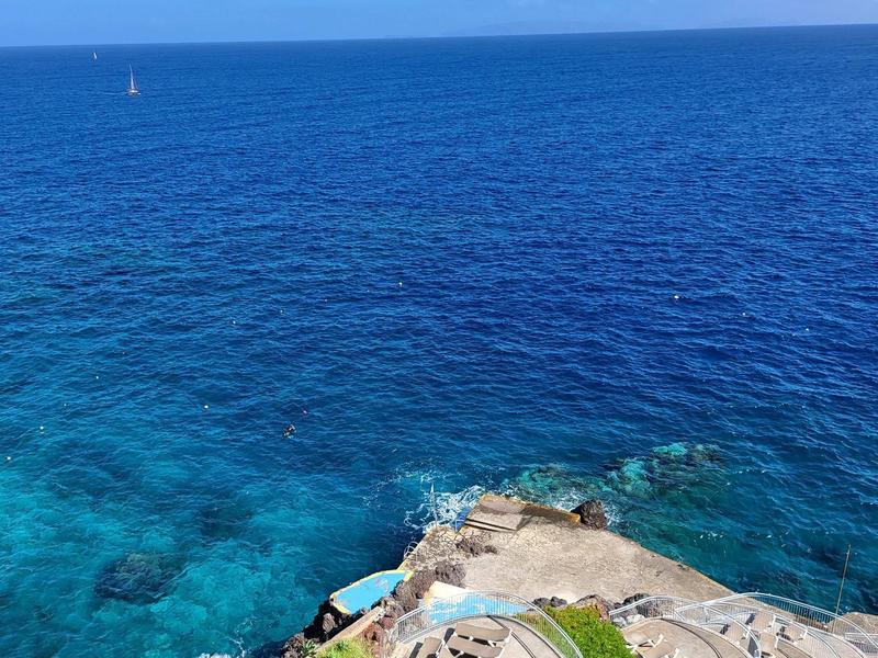 Výhled na čisté modré moře s pobřežní skalou a schody vedoucími k vodě.