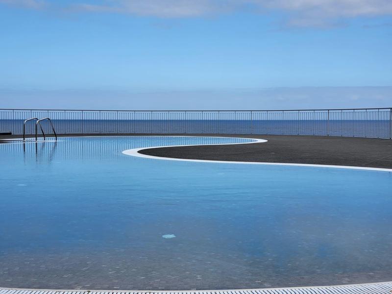 Kulatý venkovní bazén s čistou modrou vodou pod modrou oblohou a zábradlím v pozadí.