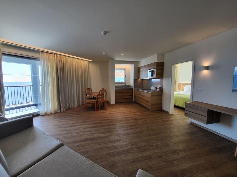 Světý hotelový pokoj s kuchyňským koutem, jídelním stolem a balkonem s výhledem na moře.