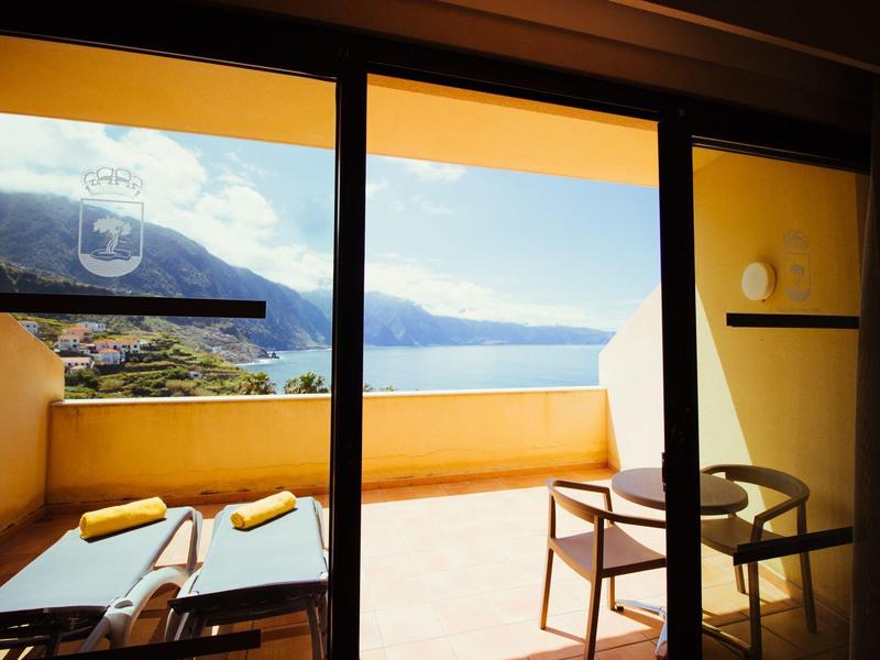 View from a balcony with lounge chairs and a table overlooking a lake and mountains on a clear day.