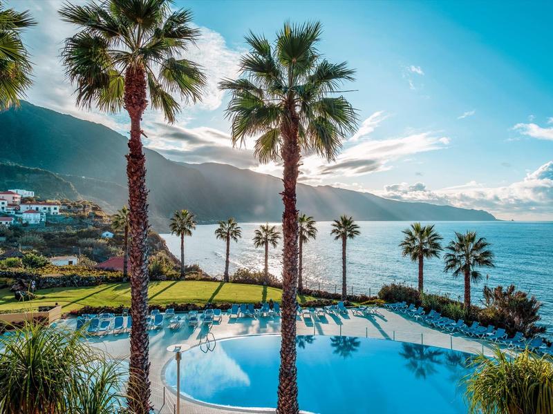 View of a pool with palm trees and sea under a clear sky at a resort.