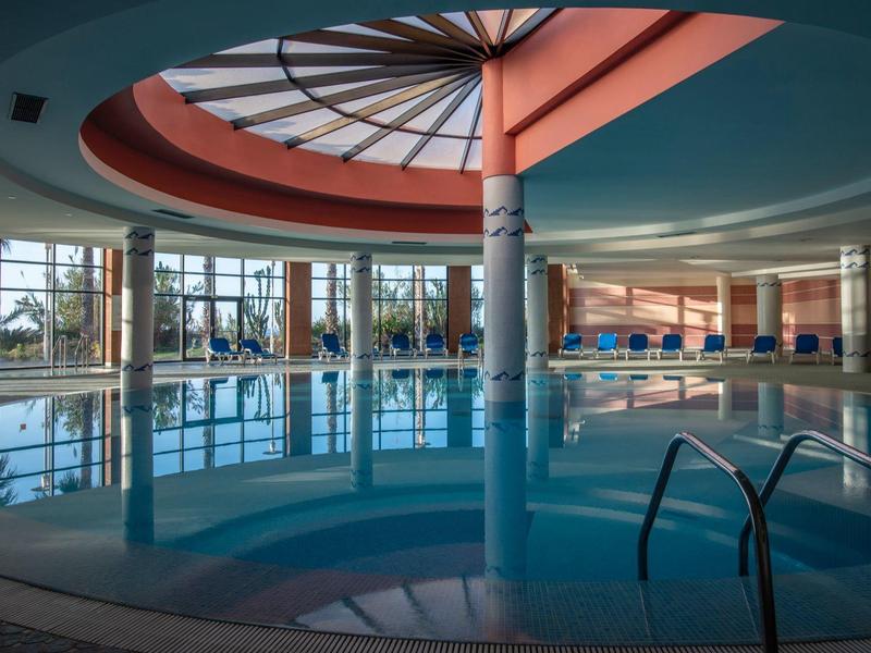 Large indoor pool with circular ceiling, windows, and water slide in a hotel.