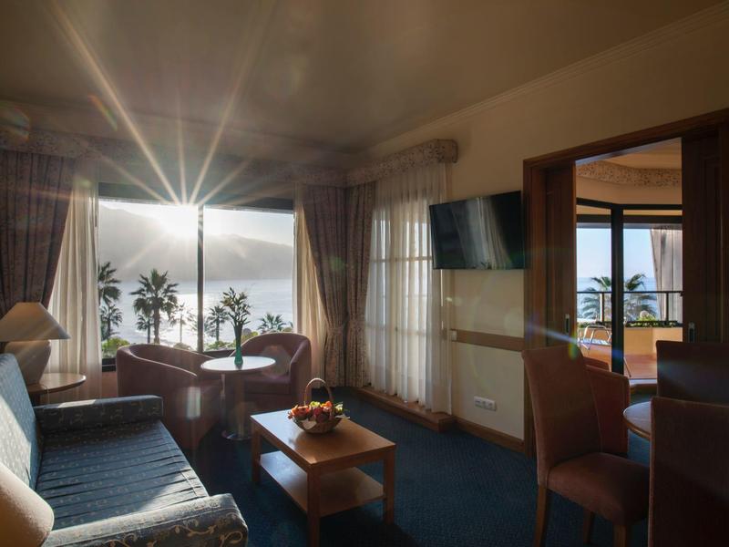 Bright hotel room with sea view, sofa, table and chairs, palm trees visible through windows.