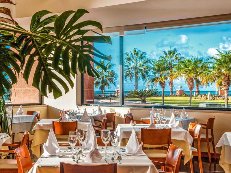 Elegant restaurant with set tables and view of palm trees and sea.
