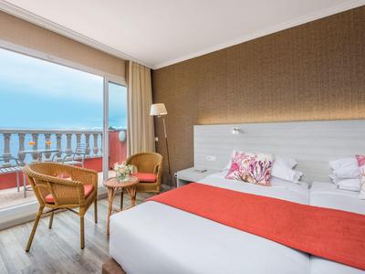 Helles Hotelzimmer mit Doppelbett, roten Akzenten, Balkon und Meerblick bei Tageslicht.