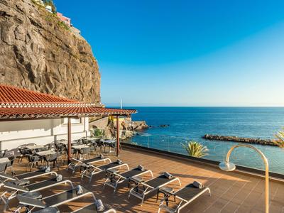 Terrasse mit Liegestühlen neben dem Meer, Felsen im Hintergrund, klarer blauer Himmel, sonnige Szenerie.