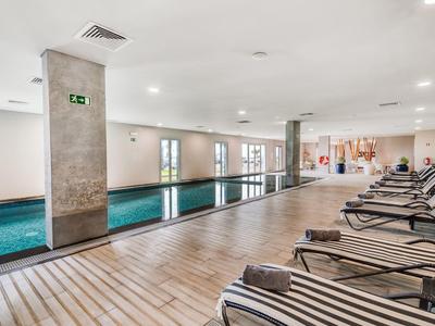 Innenraum eines Schwimmbads mit langem Becken, Holzfußboden und Liegestühlen mit gestreiften Polstern.