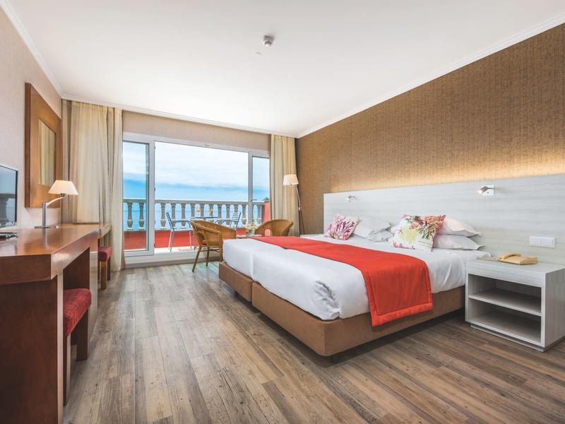 Bright hotel room with double bed, wooden floor, and balcony with sea view.