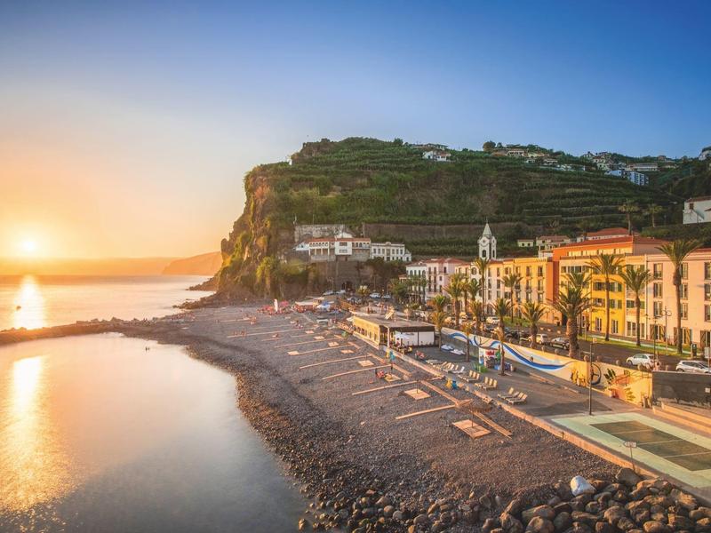 Sunset over a coastal town with harbor, beach, and colorful houses by the water.