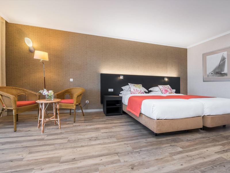 Modern hotel room with two single beds, chairs, table, and warm lighting.