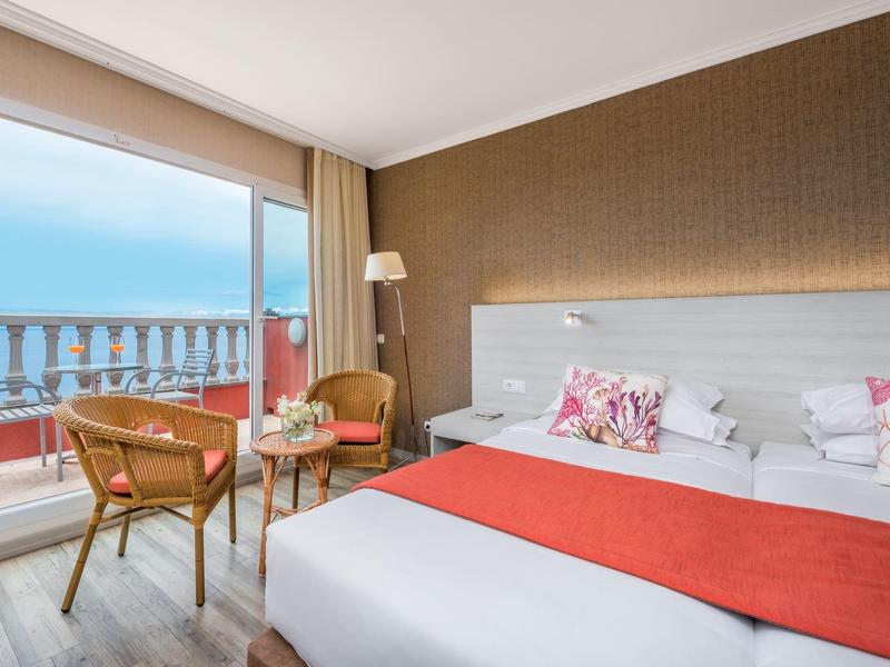 Modern hotel room with balcony and sea view, light furniture, and red accents.