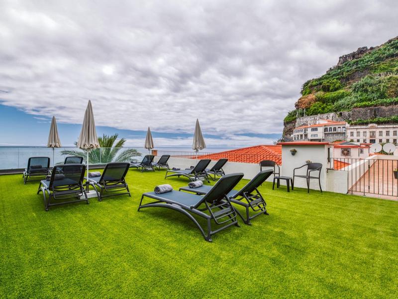 Lounge chairs and umbrellas on a rooftop terrace overlooking the sea and hills.