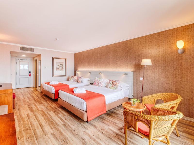 Bright hotel room with two beds, wooden floor, and seating area with chairs and table.
