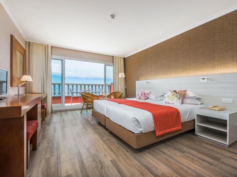 Modern hotel room with double bed, wooden floor, and balcony overlooking the sea.