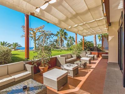 Terrassenbereich mit bequemen Polstersesseln, Tischen und Sonnensegel bei klarem Himmel.