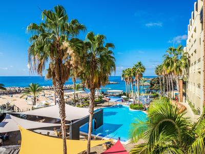 Hotelpool mit Palmen, Liegestühlen und Blick auf das blaue Mittelmeer unter klarem Himmel.