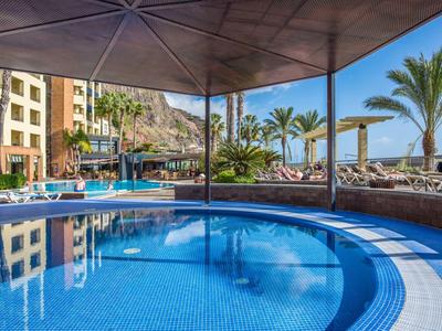 Runder Pool mit blauer Fliesen, Sonnenschirm und Palmen vor Hotelgebäuden und Bergkulisse.