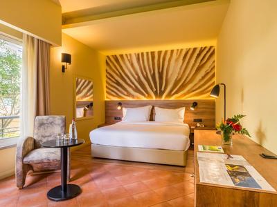 Modernes Hotelzimmer mit großem Bett, Sessel, Fenster, Holzmöbeln und dekorativem Wandbild.