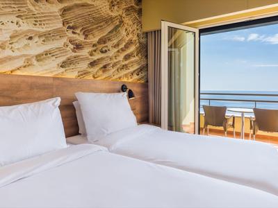 Helles Schlafzimmer mit zwei Betten, Holzwand, offener Tür und Blick auf Meer und Terrasse.