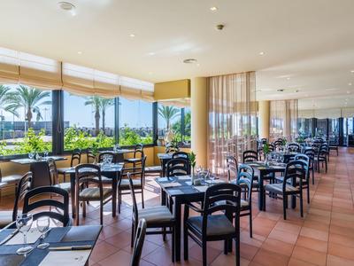 Helles Restaurant mit vielen schwarzen Stühlen und Tischen, großer Glasfront und Blick auf Palmen.