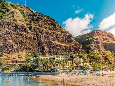Hotel am Strand mit hohen Felsen im Hintergrund und blauem Himmel bei Sonnenschein.