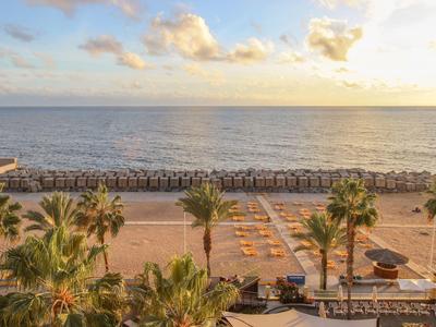 Strand mit Palmen und Liegestühlen, darüber bewölkter Himmel und ruhiges Meer im Sonnenuntergang.