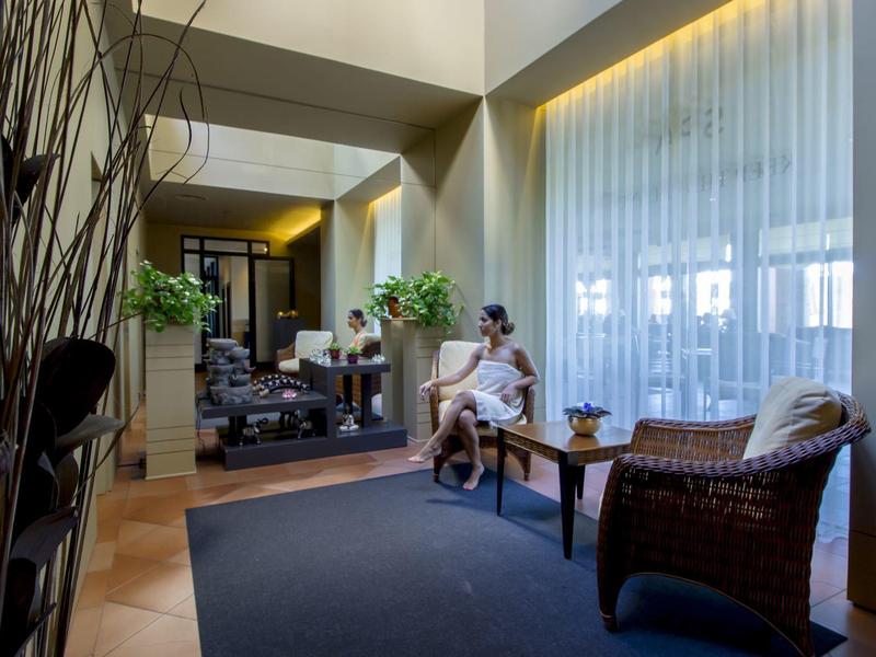 Heller Wartesaal mit Korbstühlen, kleinem Holztisch, Pflanzen und großem Spiegel an der Wand.