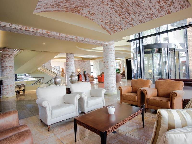 Heller Lounge-Bereich mit weißen Säulen, Ledersesseln, HolzmittelTisch und großen Fenstern.