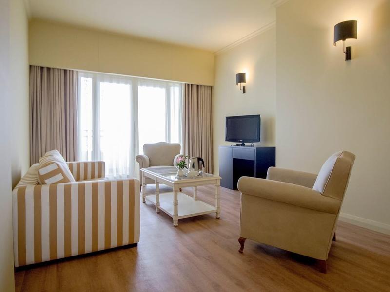 Helles Wohnzimmer mit Holzfußboden, zwei Sessel, gestreiftem Sofa, TV und großem Fenster mit Vorhängen.