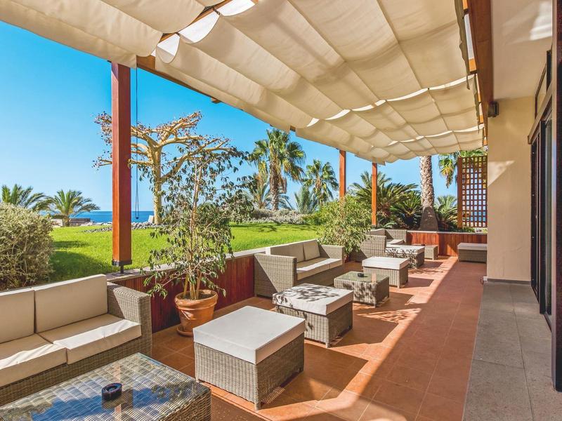 Terrassenbereich mit bequemen Polstersesseln, Tischen und Sonnensegel bei klarem Himmel.