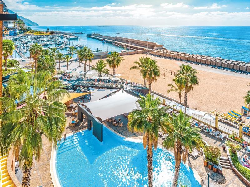 Großer Pool mit Palmen, Strand und Meer im Hintergrund, helle Sonne und klare blaue Himmel.