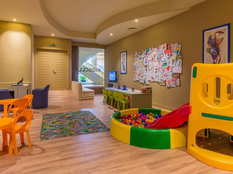 Kinderspielzimmer mit buntem Ballbecken, Rutsche, Tischen, Stühlen und Wanddekoration in warmem Licht.