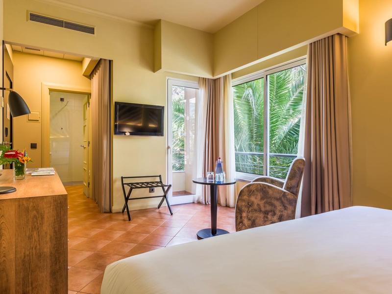 Helles Hotelzimmer mit großem Fenster, Bett, Stuhl, TV, Holzmöbeln und warmen Wandfarben.