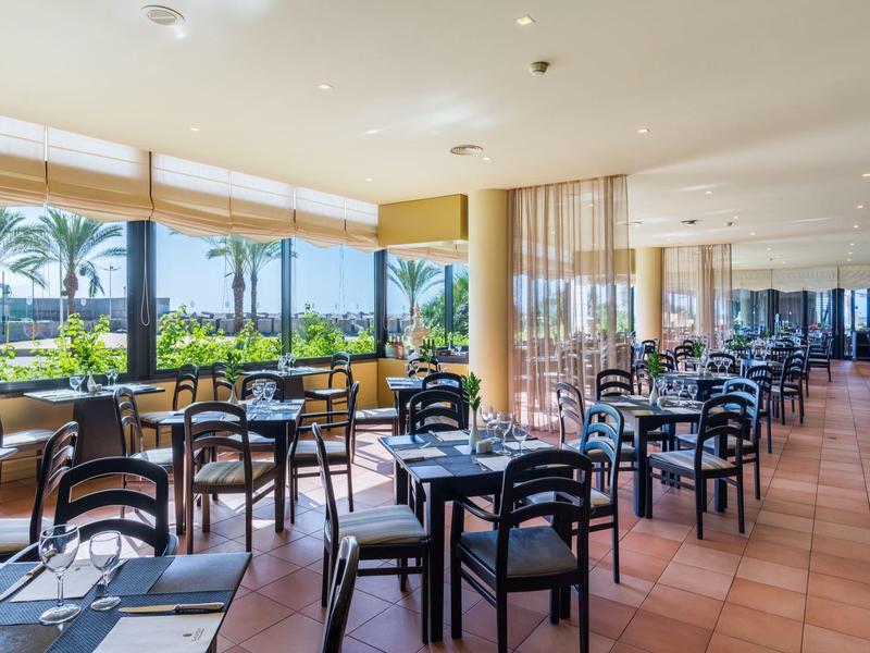 Helles Restaurant mit vielen schwarzen Stühlen und Tischen, großer Glasfront und Blick auf Palmen.