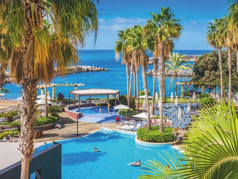 Blick auf blauen Pool mit Palmen und Meer im Hintergrund unter strahlend blauem Himmel.